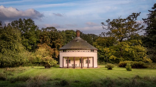The exterior of The Round House, a rotunda cottage with a portico over a patio and large, lawned garden, surrounded by trees, Suffolk